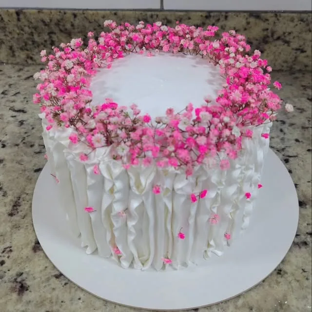 Bolo branco com babados verticais na lateral e uma coroa de flores de mosquitinho em tons de rosa no topo